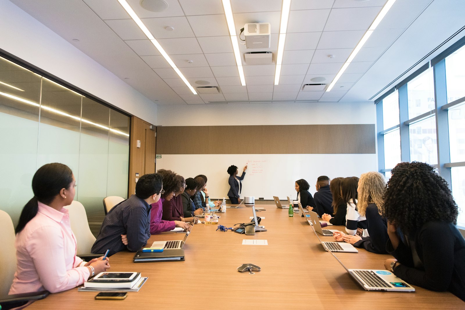 Participants around a conference table listen as a presenter writes on a whiteboard.